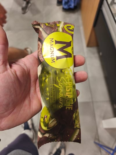 A close-up, eye-level shot captures a person's left hand holding a Magnum ice cream bar in its packaging. The ice cream wrapper is prominently featured, displaying a vibrant yellow "MAGNUM" logo and the flavor name "A-PISTACHE Signature" in gold script. Text on the wrapper further describes the product as "COATED WITH PISTACHIO. DIPPED IN MAGNUM CHOCOLATE." Additional details visible on the packaging include "CRACKING SHELLS," "257 kcal," and "90 ml/69.5g."
The background is softly blurred, indicating an indoor setting, possibly a shop or commercial establishment, with light-colored flooring and dark furniture or shelving visible. Portions of other people's legs and feet, wearing shoes, are also visible in the blurred background, suggesting movement or other patrons nearby. The focus is entirely on the hand and the ice cream, drawing attention to the product. The scene takes place in Torrent, Spain.