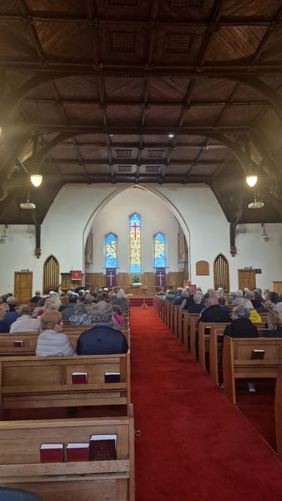 The image depicts the interior of a church in Llandudno, United Kingdom, filled with congregants seated in wooden pews. The scene is viewed from the back of the church, looking towards the altar area. The ceiling is a prominent feature, with intricate wooden paneling and beams. Pendant lights hang from the ceiling, illuminating the space.
In the foreground, wooden pews are visible, with some congregants seated. Several books, likely hymnals or Bibles, are placed in the racks of the pews. A bright red carpet runs down the center aisle.
Further down the aisle, the altar area is visible. There is a pulpit on the left, and a large, arched stained-glass window above the altar. Two banners with crosses hang on either side of the altar, and the words "PEACE" and "HEALING" are visible on them. A lectern is also present.
The walls are painted white, and there are smaller arched windows on either side of the main stained-glass window. The overall atmosphere suggests a religious service or gathering is in progress. The congregation appears to be predominantly composed of older adults. The architecture suggests a traditional church building.