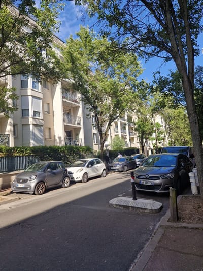 The image shows a street in Antony, France, lined with trees and a cream-colored apartment building. Several cars are parked along the street. In the foreground, a grey Smart car with license plate "FB 716 QT" is parked on the left, and a white car is parked to its right. Further down the street, a grey Citroën car with license plate "DR 290-ZB" is visible, along with a dark van and other vehicles. The trees have lush green foliage, and the sky is blue with some clouds. A metal fence with a hedge behind it runs along the front of the apartment building. The scene appears to be a typical residential street on a sunny day.