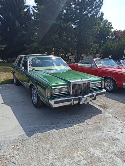 The image captures an outdoor event, likely a car show, in Mariánské Lázně, Czechia. In the foreground, a gleaming green four-door sedan, possibly a Lincoln Town Car from the late 1970s or early 1980s, is parked on a concrete surface. The car has chrome accents, a prominent grille, and a license plate that reads "1K6 7539 CZ". Behind it, a red convertible is parked, with its top down, revealing a light-colored interior.

The background features a line of mature evergreen trees, casting long shadows on the ground, indicating a sunny day. A few individuals are visible in the middle ground, some standing and conversing, others seated. Their attire suggests a casual outdoor gathering. To the left, a bicycle is leaning against something obscured by foliage. Further back, two small, shed-like structures are visible. The overall scene depicts a relaxed atmosphere at an outdoor event, likely a classic car exhibition, set against a backdrop of lush greenery.