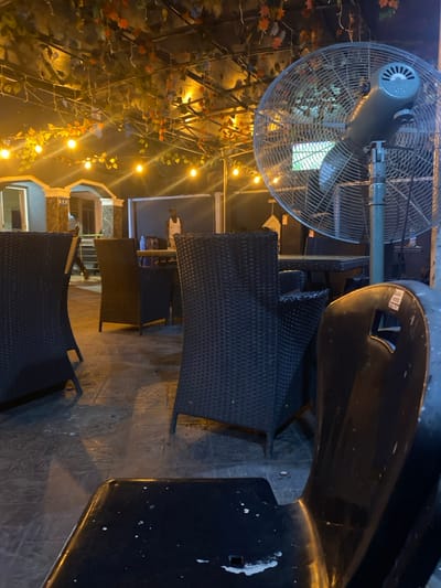 The image depicts an outdoor patio area at night in Igbessa, Nigeria. The scene is illuminated by multiple warm yellow string lights draped across a metal framework adorned with artificial green and reddish-brown foliage. The ground is paved with dark grey square pavers.
In the immediate foreground on the right, the backrest and seat of a black plastic chair are visible, showing wear with white splatters and a partially discernible "TYSON" label. To the right, a large grey industrial-style standing fan with a circular cage is positioned.
The midground features multiple dark brown or black wicker-style chairs and tables arranged on the patio. In the central background, a male figure wearing a white tank top stands near one of these tables. To the left background, a blurred figure is visible near the entrance of a building with light-colored columns and arches. A dark wall or fence forms the distant background boundary of the patio. No direct interactions between individuals are evident.
