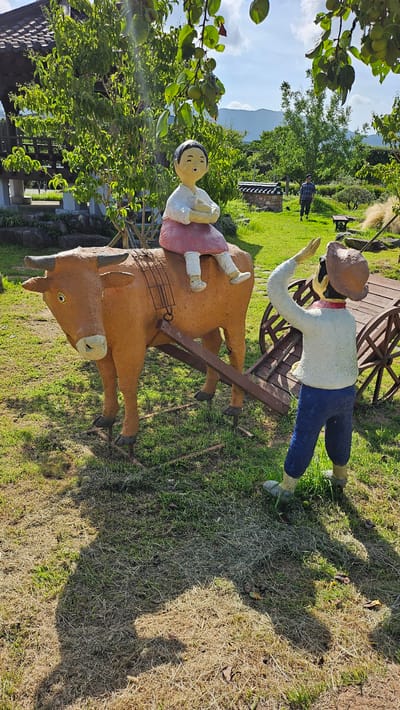 This outdoor image depicts a scenic view with sculptures in the foreground and a natural landscape in the background. The sculptures represent a farmer guiding a bull, with a child seated on the bull. The farmer, dressed in a white shirt and blue pants, is facing away from the viewer, with his right arm raised as if to direct or communicate. The bull, painted in an earthy brown hue, is harnessed to a wooden cart, with the child, dressed in a pink skirt and white top, seated atop the bull. The scene is set in a grassy area with some dry patches and fallen leaves, suggesting a park or rural environment. In the background, there is a traditional Korean tiled roof structure, lush green trees, and mountains in the distance under a clear blue sky with scattered clouds. A person in casual attire can be seen walking in the middle ground. This scene is located in Geoje-si, South Korea, likely at a cultural or historical attraction that showcases traditional Korean life.