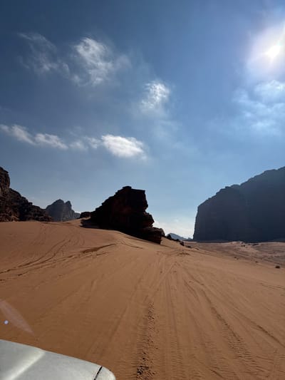 The image depicts a desert landscape in Ash Shākirīyah, Jordan, captured from a light-colored vehicle whose front left portion is visible in the lower foreground. The scene features extensive reddish-orange sand dunes crisscrossed by numerous vehicle tire tracks. Dominating the environment are large, dark, weathered sandstone rock formations: a range of reddish-brown mountains extending from the left, a prominent dark freestanding formation in the midground, and a massive, steep dark mountain face on the right horizon. The sky is clear blue with scattered white clouds, and the sun, positioned in the upper right, generates a visible lens flare. No individuals are present.