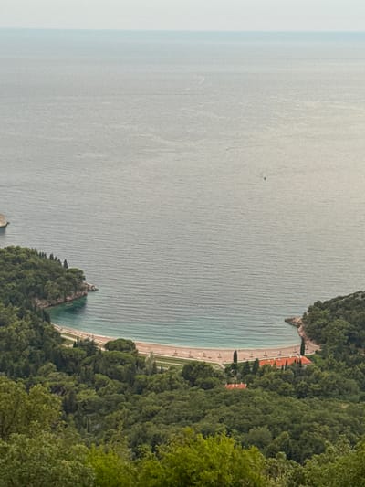 The aerial view captures a crescent-shaped beach nestled within a lush, green cove. The sandy beach is lined with a path and several small buildings with orange tile roofs. The turquoise water of the Adriatic Sea gently laps the shore, with a few distant boats visible on the horizon. The scene is dominated by dense, green foliage, with trees cascading down the hillsides that surround the bay. The overall impression is one of a secluded and tranquil coastal landscape in the vicinity of Čelobrdo, Montenegro.