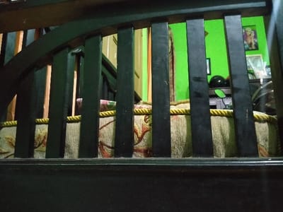 The image is a close-up view of the back of a wooden bench with a padded seat. The bench is located in a home in Lagadar, Indonesia, at the coordinates -6.928 latitude and 107.535 longitude. The bench is made of dark wood and has a high back with vertical slats. The seat is covered in a patterned fabric with a yellow trim. The background is blurred and shows a green wall with a door and a picture frame. There are other objects in the background that are too blurry to identify. The scene appears to be taken from a low angle, as the viewer is looking up at the back of the bench. The bench is empty. There are no people present in the image.