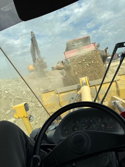 The image, taken from inside the cab of a heavy machine in Hristiyanovo, Bulgaria, looks out through a dusty windshield onto an active quarry or construction site. In the immediate foreground, a black steering wheel with the "Komatsu" logo on its horn pad is prominent on the right. Behind the steering wheel, a dashboard is visible, featuring a speedometer reaching up to 40 km/h, along with multiple gauges (fuel, temperature, etc.) and various warning lights. Part of a person's leg, clad in dark pants, is visible at the bottom right, suggesting an operator is seated in the cab.

Outside the vehicle, a large-scale rock processing operation is underway. Directly ahead, a substantial yellow construction machine, likely a wheel loader or excavator, has its arm and bucket partially visible. To the left of this machine, a large red and gray crushing or screening plant is actively processing rocky material, generating a significant amount of dust that hangs in the air. Piles of freshly processed or raw aggregate are visible around the plant. Further in the background, another large yellow piece of equipment, possibly an excavator or drilling rig, extends its arm against the sky.

The sky in the background is bright and partly cloudy, indicating daytime. The entire scene is characterized by industrial activity and dusty conditions typical of a quarry or large construction site.