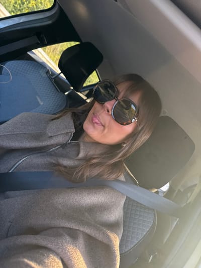 A woman is captured in a selfie taken inside a car. She is positioned in the driver's or front passenger's seat, with a light-colored car ceiling visible above her and dark headrests behind her. She has medium-length, dark brown hair with bangs and is wearing oversized, dark-rimmed sunglasses, possibly with a tortoiseshell pattern. Her head is tilted slightly, and she is playfully sticking out her tongue. A small, gold-colored hoop earring is visible on her right earlobe. She is dressed in a thick, light brown or taupe-colored coat with a prominent silver zipper down the front, indicating a warm, wool-like material. A light grey seatbelt is fastened across her chest.
The car's interior features grey seats with a textured, possibly quilted, diamond pattern visible on the headrests and seat upholstery. Through the car windows on the left side of the image, green foliage and what appears to be a road barrier or railing can be seen, suggesting the car is either parked by a road or in motion. The overall lighting is bright and sunny, casting a warm glow on her coat and creating reflections on her sunglasses, indicating daytime, likely morning or late afternoon due to the angle of the sunlight. No text is visible in the image.