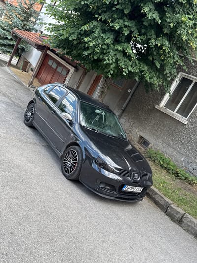 This image shows a black Seat Leon parked on a paved surface, likely a street or driveway, in Dobrinishte, Bulgaria. The car is angled, with its front slightly towards the right side of the frame. It has aftermarket alloy wheels with a multi-spoke design and red accents on the brake calipers. The license plate is visible, reading "BP5871CX".

To the left of the car, there is a building with textured gray walls and a dark-tiled roof. A large tree with dense green foliage overhangs the car and the building. The tree branches obscure some of the building's details, including a window. A portion of a wooden door and a smaller window are also visible on the building's facade.

The ground is a rough, gray asphalt. A concrete curb runs along the edge of the paved area, showing signs of cracking. Behind the curb, there is an area of dry grass and some greenery. The lighting suggests it is daytime, possibly late morning or early afternoon, with bright sunlight creating reflections on the car's surface. The overall scene depicts a residential or semi-rural setting. No people are visible in the image.