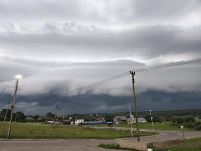 This image shows a rural scene in Ateptsevo, Russia, under a dramatic, stormy sky. The sky is filled with dark, ominous clouds that suggest an approaching storm, with a band of lighter, layered clouds stretched across the horizon. In the foreground, a paved road curves to the right, bordered by green grass and wildflowers. Several utility poles with wires are visible, including one with a lit lamp on the left. In the middle ground, a cluster of houses and other buildings forms a small village. Trees and greenery surround the village, with a dense forest forming the backdrop. A few signs, including a yield sign, are visible on the side of the road. There are no people or vehicles prominently featured in the image. The overall atmosphere is one of impending weather change in a quiet, rural setting.