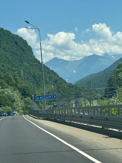 The image shows a view from a car on a highway, with a mountainous landscape on either side. In the distance, snow-capped mountains are visible under a blue sky with white clouds. The road curves to the left, and a metal guardrail lines the right side of the highway. A sign with blue text that reads "р. МЗЫМТА" (R. Mzymta) is visible on the left side of the road, near a lamppost. The area appears to be Chvizhepse, Russia, a resort town in Krasnodar Krai, known for its natural beauty and mountainous terrain. The scene depicts a typical scenic drive through a mountainous region.