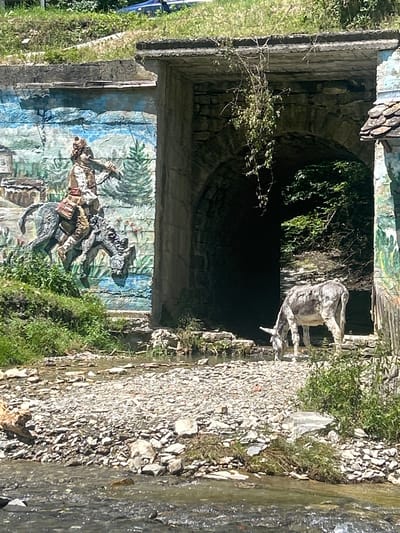 In the city of Beli Osam, Bulgaria, a gray donkey with black markings is grazing near a stone tunnel entrance. The tunnel is part of a stone structure, possibly a bridge or viaduct, with rough-hewn stone blocks forming the archway. The tunnel is dark inside, with lush green foliage visible at the back. To the left of the tunnel entrance, a colorful mural is painted on a stone wall. The mural depicts a figure in historical attire riding a horse, with a stylized landscape of trees and buildings in the background. The ground in front of the tunnel is a mixture of rocks and pebbles, with a shallow stream flowing through it. Green grass and bushes grow around the area, and some dead branches hang from the tunnel's arch. The scene is illuminated by daylight, casting shadows.