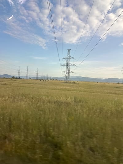 This is a landscape photograph taken from a moving vehicle, likely a train or car, looking out over a grassy field and a line of electrical pylons under a cloudy sky. The image is oriented vertically, with the horizon running from top to bottom along the right side of the frame.
The foreground is dominated by a blurry, green expanse of grass, indicating movement. Several tall, lattice-style electrical pylons are visible in the mid-ground, marching in a line towards the background. They are silhouetted against a pale blue sky scattered with white, puffy clouds. In the far background, distant mountains or hills are visible, their forms softened by the atmospheric haze.
The lighting suggests it is daytime, with the sun filtering through the clouds. The overall impression is one of a rural or semi-rural landscape, typical of open country. There are no people or specific man-made structures visible other than the electrical infrastructure. The dynamic blur of the grass emphasizes the perspective of being in transit, experiencing the scenery as it passes by. There is no visible text in the image.