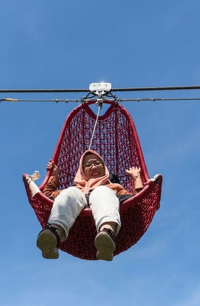 A woman, wearing a hijab and glasses, sits in a large, red, woven pod-shaped swing suspended from a wire against a clear blue sky. She is smiling and waving, with her legs dangling down. Her outfit consists of a light brown jacket over a dark top, and loose white pants. She wears brown sneakers. The swing is attached to a metal pulley system that runs along the wire. The sun is bright, suggesting it is daytime, and the weather is clear. The scene appears to be an outdoor recreational area, possibly an adventure park, given the nature of the swing and the overhead cables. The location is identified as Bintang Meriah, Indonesia. There is some text visible on the metal pulley mechanism, but it is too small and blurry to be read.