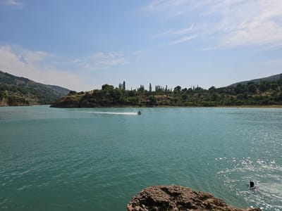 The image depicts a serene natural landscape on a bright, sunny day. The scene is dominated by a large body of calm, turquoise water, likely a lake or reservoir, reflecting the clear blue sky dotted with wispy white clouds. Lush green hills covered in trees and vegetation surround the water, creating a picturesque backdrop.
In the middle of the water, a person is riding a jet ski, leaving a white wake behind them, indicating movement and recreational activity. Further to the right, closer to the foreground, another person is visible in the water, with their head and shoulders above the surface, suggesting they are swimming or wading.
The time of day appears to be midday, given the brightness of the sunlight and the shadows cast. The weather seems pleasant and warm, conducive to outdoor water activities. The landscape, with its hills and vegetation, points to a rural or semi-natural location. The presence of both a jet ski and a swimmer implies this is a popular spot for leisure and water sports. The overall mood conveyed is one of tranquility and enjoyment of nature's beauty and recreational opportunities. There is no visible text in the image.