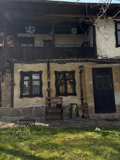 The image captures a building facade in the village of Gorno Trape, Bulgaria. The structure exhibits an old-world charm with its stucco walls, dark wood trim, and aged stone foundation. Two distinct windows with uniquely shaped dark wood frames punctuate the wall, reflecting the outdoor scenery. Above the windows, a dark wood door provides access to the building, while below, a wooden chair and various tools and containers are placed against the wall. To the left of the building, a partial view of a timber-framed structure is visible, adorned with a hanging floral arrangement. On the right, a stone pathway transitions into a grassy area with scattered white flowers, creating a contrast between the man-made structure and the natural environment.