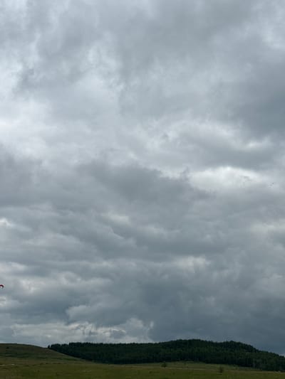 The image shows a cloudy sky over a rural landscape. In the distance, there is a treeline atop a hill, and in the foreground, a grassy field. A small red object, possibly a kite or part of one, is visible in the upper left portion of the image, low in the sky. The scene appears to be outdoors, with no visible buildings or infrastructure immediately apparent. The location is identified as Staroye Subkhankulovo, Russia.