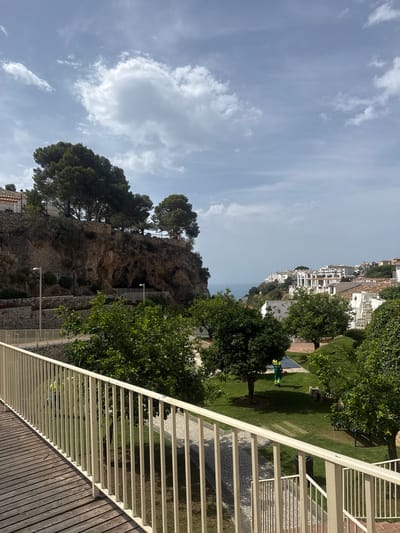 The image depicts a view from a wooden walkway with a white railing, looking towards a cliffside with trees and buildings. In the foreground, the walkway runs diagonally from the bottom left. Behind the walkway, a lush green garden area is visible, with several trees. In the garden, a person wearing a yellow vest and green pants is operating a leaf blower. Beyond the garden, a steep, rocky cliff face rises, topped with pine trees and a few buildings. The cliff has a dark opening, possibly a cave or an overhang. Further in the background, a white-washed town is nestled on a hillside overlooking the sea, which is visible on the horizon. The sky is partly cloudy with a mix of blue sky and white clouds. The architecture of the town in the background, with its white buildings and tiled roofs, is characteristic of coastal towns in southern Spain, and likely Benalmádena, Spain, given its hilly terrain and proximity to the sea.
