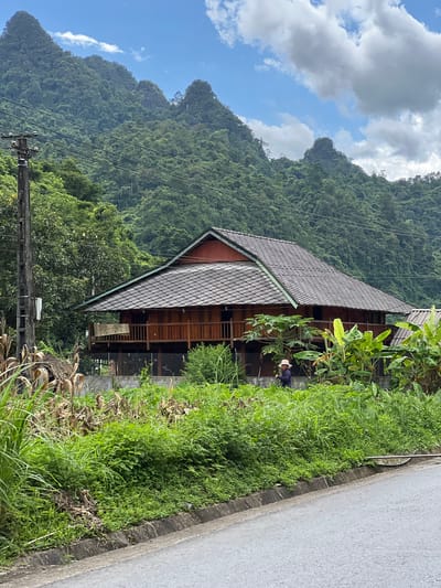 The image shows a traditional wooden stilt house with a tiled roof, set against a backdrop of lush green mountains and a partly cloudy sky. The house features a wide wooden balcony with railings. In front of the house, there is a person wearing a hat and a dark colored shirt, who appears to be tending to the vegetation. The foreground consists of a paved road, a concrete curb, and an area filled with various plants, including tall grasses and what appear to be corn stalks. A utility pole with wires is visible to the left of the house. The scene is characteristic of a rural setting in Thuận Hòa Commune, Vietnam, emphasizing a harmonious blend of traditional architecture and the natural landscape.