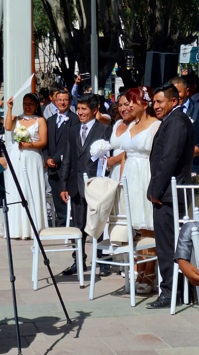 A wedding party is posing for a photograph, seated in rows on white chairs. The scene unfolds outdoors. The presence of trees suggests the location to be Cerro Colorado, Peru. The party consists of several couples dressed in wedding attire: the women in white dresses and the men in black suits. A photographer is visible with camera equipment.