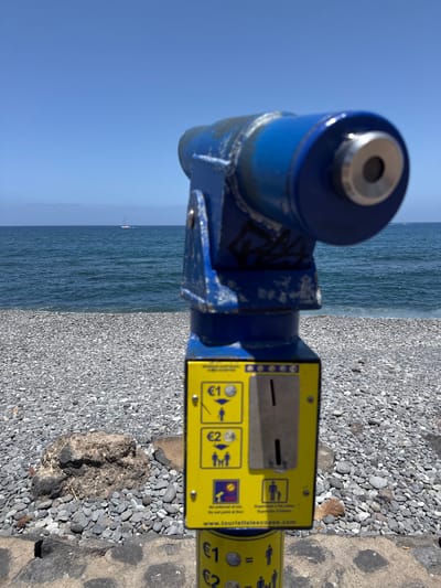 A blue coin-operated tourist telescope is mounted on a blue metal stand, set against a backdrop of the sea and a pebble beach. The telescope is positioned to the left of the frame, with its lens pointing towards the ocean. To the right of the telescope is a yellow payment panel with slots for coins and instructions in English and other languages. Text on the panel includes "www.touristtelescopes.com" and mentions options for €1 and €2. The sea is calm and blue, with a small sailboat visible in the distance. The beach is covered in smooth, grey pebbles. The sky is clear and bright blue, suggesting a sunny day. The scene appears to be set at a scenic coastal location, possibly a promenade or viewpoint. The image is vertically oriented.
