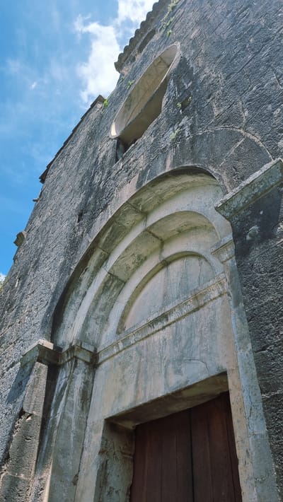 This is a low-angle, close-up, exterior shot of a stone structure in Sales de Llierca, Spain. The stone is dark gray and appears weathered, with some areas showing lighter green moss or lichen. The image is rotated so that the sky, which is a vibrant blue with scattered white clouds, occupies the lower portion of the frame.
The structure features an arched entryway, detailed with concentric curved stone moldings. To the left of this arch, a smaller, oval-shaped opening is visible in the stone wall, possibly a window or niche. The upper right portion of the image shows a section of a wooden door. The stone around the doorway is lighter in color and shows signs of age, with some cracking and discoloration.
The lighting suggests it is daytime, with bright sunlight illuminating the scene. The overall impression is of an ancient, possibly historical, building. The detailed stonework and the setting against the sky hint at an architectural feature of a church, castle, or similar edifice. There is no visible text in the image.