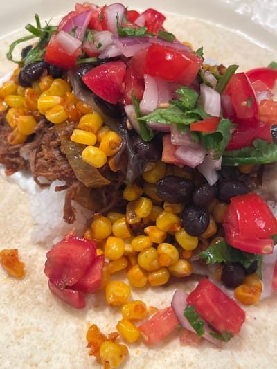The image displays a detailed close-up of a prepared food item, consistent with a generously filled burrito or large taco, presented on a light-colored tortilla. The central focus is a mound of layered ingredients, including a base of white, granular material, likely rice, overlaid with shredded, reddish-brown meat. This is mixed with and topped by bright yellow corn kernels, whole black beans, and translucent cooked onion segments. The uppermost layer consists of a fresh topping composed of diced red tomatoes, diced red onions, and chopped green herbs, consistent with cilantro. Several individual corn kernels and diced tomato pieces are scattered onto the surrounding tortilla surface. The immediate background is a plain, light surface, likely a plate. No visual indicators are present within the image to determine its geographic location, including a location in Merom HaGalil Regional Council, Israel.