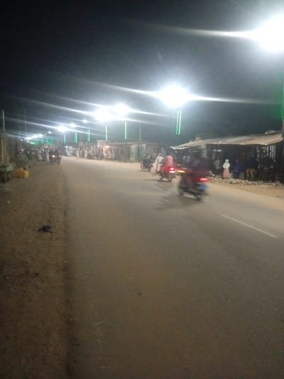 This media file captures a nighttime scene on a street in Tundun Wada West, Nigeria. The image is dominated by the bright glare of streetlights, suggesting artificial illumination. The road surface appears to be asphalt, with a dirt and gravel shoulder on the left side. Along the right side of the road, there are several makeshift structures, possibly market stalls or small shops, with corrugated metal roofs.

Several people are visible, some standing and others on motorcycles. The individuals appear to be dressed in casual clothing and some women are wearing headscarves. The presence of multiple motorcycles, some in motion and blurred due to the long exposure, indicates that motorbikes are a common mode of transportation in this area. One motorcycle in the foreground appears to be moving away from the camera, with its taillight illuminated.

The overall atmosphere suggests a lively evening, possibly a marketplace or a busy thoroughfare at night. The lighting creates a dramatic effect, casting long shadows and highlighting the dust and texture of the road. There is no visible text in the image. The weather appears to be clear, given the absence of rain or fog. The scene conveys a sense of everyday life and activity in a Nigerian community after dark.