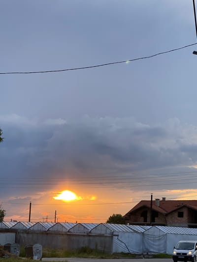 A vibrant sunset illuminates a rural or semi-rural scene in Miriantsi, Bulgaria, captured during dusk. The image is dominated by a dramatic sky, split between deep blue-gray on the left and intense orange and yellow hues on the right, where the sun glows brightly through and behind clouds. The sun itself is a powerful, diffuse light source, partially obscured by thick, dark clouds that reflect its warm colors.

Several dark electrical wires stretch horizontally across the upper and middle sections of the frame, contrasting against the colorful sky. A faint, small green-yellow light or reflection is visible on one of these wires towards the upper left. On the far right, a two-story brick house with a red tiled roof appears to be under construction or unfinished, featuring dark, empty window openings and a prominent chimney. Adjacent to and in front of the house are multiple structures resembling greenhouses or covered sheds, characterized by concrete block bases and translucent white, sloped coverings. These structures extend across the middle-right of the image, below the house. A utility pole is also visible amidst these structures.

A white van is partially visible parked on the far upper right, with a license plate displaying "B 544AX" (partially legible). In the lower right corner, two metallic garbage cans or large bins are situated near the edge of the property. Sparse vegetation, including trees and grass, can be seen near the buildings and along the ground. No human activity is depicted, focusing instead on the atmospheric landscape and the interplay of natural light with man-made elements.