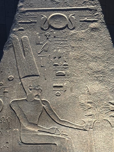 This is a close-up, vertical, eye-level photograph of a relief carving on a large stone slab, likely part of an ancient Egyptian monument. The carving depicts a seated figure wearing a tall, segmented headdress and intricate layered jewelry. The figure's right arm is extended, and their hand appears to be grasping or interacting with something beyond the frame of the image to the right. Hieroglyphs are carved into the stone above and to the left of the figure. The stone surface is textured and weathered, showing signs of age. The lighting creates strong shadows, highlighting the relief work and the granular texture of the stone. The background appears to be a dark, textured surface, possibly a wall or another stone structure. The relief is located in the city of Kafrat Nassar, Egypt.