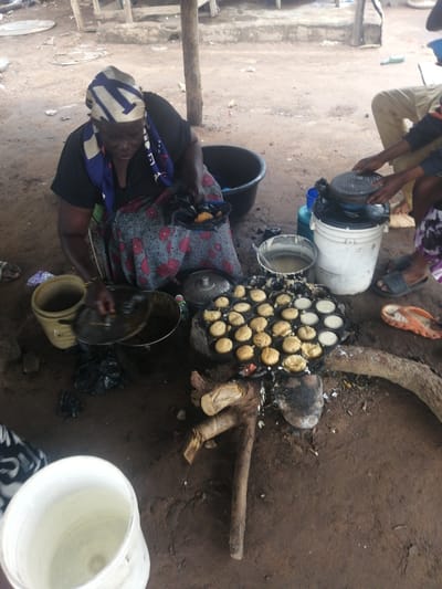 The image captures a scene of food preparation at a market stall in Ajasse Ipo, Nigeria. In the foreground, a woman wearing a black top and a patterned skirt is seated and actively engaged in cooking. She has a colorful headscarf tied around her head. In front of her is a specialty cooking pan with multiple circular molds, filled with small, golden-brown, pancake-like items. Some molds appear to be empty, suggesting the cooking process is ongoing. To her left, a yellow bucket is visible.

To the right of the woman, a person's legs are visible, wearing khaki trousers and sandals, reaching towards a white bucket. Nearby, a black plastic tub and a blue container are present. The cooking setup involves a fire pit with burning embers beneath the pan, indicating an outdoor cooking environment. The ground is dirt, typical of a market setting, with scattered debris. The background shows a rustic structure made of wood and possibly metal, with wooden posts and beams. Another person, partially visible in the background, is wearing khaki trousers and appears to be a customer or vendor. The overall impression is a bustling, open-air market where traditional cooking methods are being employed.