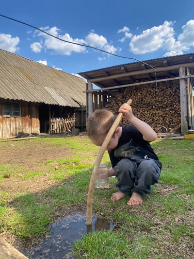 A barefoot child is squatting by a puddle, holding a stick in their hands. They are wearing a black t-shirt and grey sweatpants. Behind the child is a stack of firewood and an old wooden building with a corrugated metal roof. The sky is blue with white clouds. The ground is a mix of grass and dirt. The location is consistent with the rural characteristics of Узяр, Russia.
