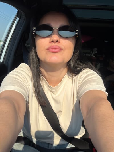 A woman, with long dark hair and sunglasses, takes a selfie while sitting in the driver's seat of a car. She is wearing a light-colored t-shirt and a seatbelt. The sun is shining brightly, casting shadows on her face and arm. The interior of the car is dark, with the sunroof open. A person is visible in the passenger seat in the background. The image does not provide enough information to determine the location within the city of Bolshiye Dvory, Russia.