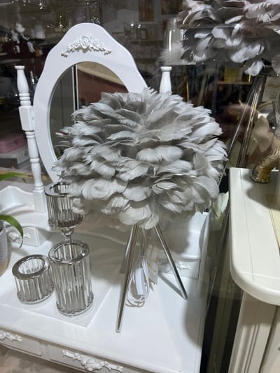 The scene depicts an illuminated retail display within a commercial establishment in the Smederevska Palanka Municipality, Serbia. A white, ornately carved dressing table or console serves as the primary surface for the merchandise. Centrally positioned on this table is a spherical table lamp featuring a lampshade composed entirely of numerous light gray feathers, supported by three slender, chrome-finished legs. A white power cord with an inline switch is visible extending from the lamp's base. To the left of the feather lamp, a freestanding oval mirror with a white, carved frame adorned with rose motifs at the top reflects a blurred interior environment. Further to the left, three silver-toned, cylindrical decorative holders, designed with vertical clear elements, are arranged on the table; one is taller and pedestal-mounted, while two are shorter. A small segment of a potted plant housed in a two-tone woven container is visible in the lower-left corner. The reflections on an apparent glass storefront or display case reveal indistinct elements of an exterior environment and additional interior merchandise. No individuals are present in the image.