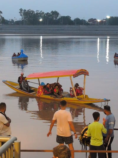The image captures a serene evening scene at the Tawi Riverfront in Jammu, India. The foreground features a railing with a light yellow banister. To the left, a man with dark hair and a white collared shirt is seen from the back, his arms crossed. To the right, a group of three men are standing near the railing. The man on the far right wears a blue t-shirt with white stripes on the sleeves and black pants. The middle man wears a bright green t-shirt and dark pants. The man on the left is wearing a white t-shirt and patterned shorts, with his back to the camera, his right arm extended slightly.
In the middle ground, two boats are visible on the calm, reflective waters of the Tawi River. The larger boat, in the center, is a yellow boat with a red canopy supported by wooden poles. It carries several passengers wearing life jackets. A person in a dark vest and a pink cap is at the stern, seemingly rowing the boat. Another smaller blue boat, shaped like a swan, is further in the distance, also carrying passengers.
The background shows a tree-lined embankment with lights illuminating the area. The sky is a soft twilight blue. The overall atmosphere is peaceful, suggesting a leisurely outing at the riverfront in Jammu.