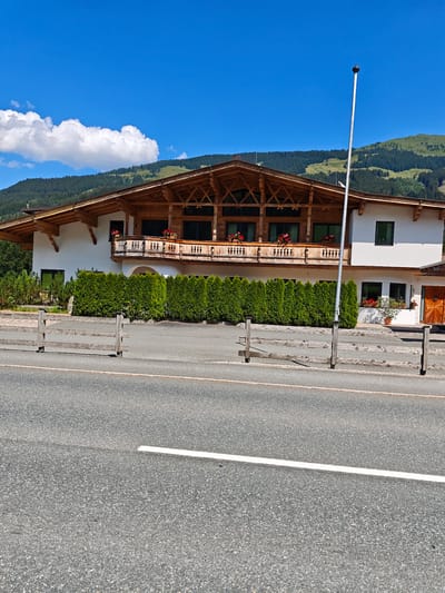 This image shows a traditional Austrian building in Filzen, Austria, on a sunny day. The building is a multi-story structure with a distinctive wooden eaves and a large balcony on the upper level, adorned with flower boxes. The facade is painted white, and there are several dark-framed windows. In front of the building is a neatly trimmed green hedge, and a rustic wooden fence runs along the foreground. A tall, thin pole stands near the center of the image. Beyond the building, rolling green hills and a forest are visible under a bright blue sky with scattered white clouds. The scene appears to be situated near a road, indicated by the asphalt and white line in the foreground. There are no people or visible activities in the image.