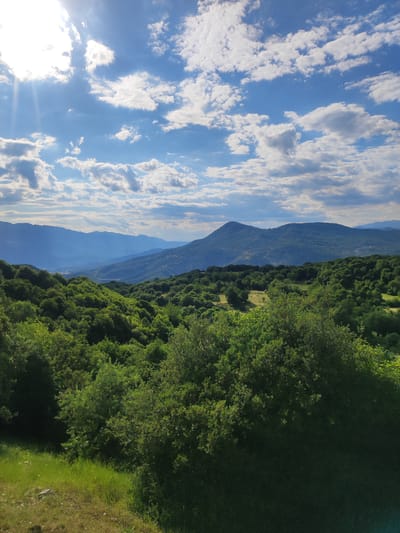 The image captures a scenic view of rolling green hills and distant blue mountains under a bright, partly cloudy sky. Sunlight beams down from the upper left corner, casting a bright glow and creating lens flare effects. The foreground is dominated by lush green foliage, with a patch of sunlit grass on the lower left. The midground features a dense forest of trees, transitioning to more sparsely vegetated hills and then to successive layers of hazy blue mountains in the background. The sky is a vibrant blue, dotted with fluffy white and gray clouds. The scene appears to be a natural landscape, likely in a rural or mountainous area, characteristic of regions like Egnatia Municipal Unit, Greece, known for its picturesque natural beauty. There are no people or man-made structures clearly visible in the image, suggesting a focus on the natural environment.