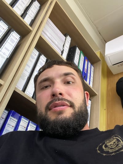 A close-up, low-angle selfie shows a man with dark hair and a beard looking at the camera. He is wearing a black t-shirt with a circular emblem of a skull and weights. He is positioned in front of a wooden bookshelf filled with binders. The background shows an air conditioning unit mounted on a yellow wall, and the ceiling has some signs of wear. The man is identified as being in Luzhitsy, Russia.