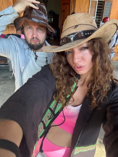 A selfie captures a man and a woman in what appears to be Bắc Hà, Vietnam, judging by the traditional clothing trim visible on the woman's jacket. The woman is wearing a tan cowboy hat with a brown band and a silver steer head adornment, a black jacket with an embroidered green trim, and a pink top. The man wears a weathered brown leather cowboy hat and a light gray button-down shirt. The background shows parts of a wooden building with barred windows and other indistinct objects.