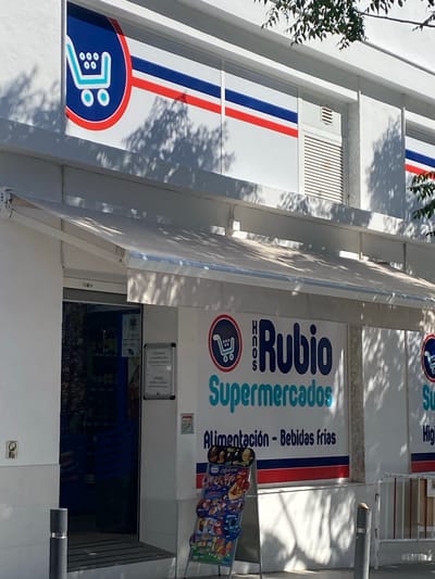 A storefront of a supermarket named "Rubio Supermercados" is visible. The building is white with a blue and red striped awning. The supermarket's logo, a blue shopping cart with four circles inside, is displayed on the facade. Below the logo, the text "Rubio Supermercados" is written in large blue letters. Underneath this, "Alimentación - Bebidas Frias" is written in smaller blue text. A display stand with various snacks and products is placed in front of the entrance. The image appears to be taken on a sunny day, with shadows of trees cast on the building. The location is identified as Conil de la Frontera, Spain.