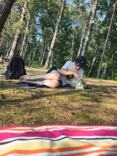 A person wearing a white t-shirt, black shorts, and a black baseball cap is lying on a blue towel on the ground in a forest. They are reaching into a bag of snacks. The ground is covered with pine needles and moss. A black backpack is situated to the left of the person, and a picnic table is visible in the background. The trees are tall and thin, with green foliage. The sun is shining, casting shadows on the ground, suggesting it is daytime. The overall mood appears relaxed and peaceful. The snack bag has the text "Xtra" visible on it.