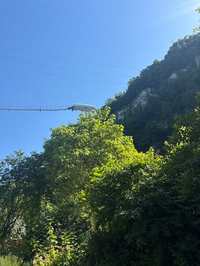 The image shows a view of a suspension bridge against a clear blue sky. The bridge stretches across a gorge, with a steep, tree-covered cliff face visible on the right. The bridge itself appears to be made of metal and has a walkway with railings. In the foreground, there are dense trees and foliage. The scene is bathed in bright sunlight, casting shadows. The location is identified as Galitsyno, Russia. There are no people visible in the image.