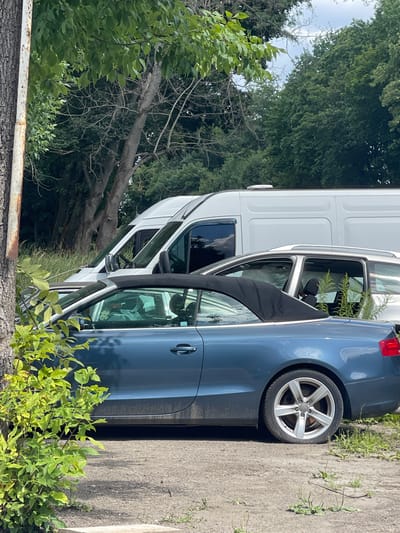 In Konopnytsia, Ukraine, on a bright day, a blue convertible car is parked on a dirt and gravel surface near a tree. The car's black top is down, and a person is visible in the driver's seat. Behind the convertible, a silver SUV is parked with its doors open, revealing vegetation growing inside. Further back, a white van is parked, with its front end obscured by the other vehicles and trees. The scene is surrounded by dense green foliage and trees.