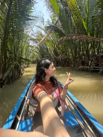 A person is in a boat floating on a river surrounded by dense, green vegetation, potentially located in Tân Thạch, Vietnam. The water is murky and brown. The boat is blue. The person has dark hair and is wearing an orange t-shirt. The person is extending fingers in the air.