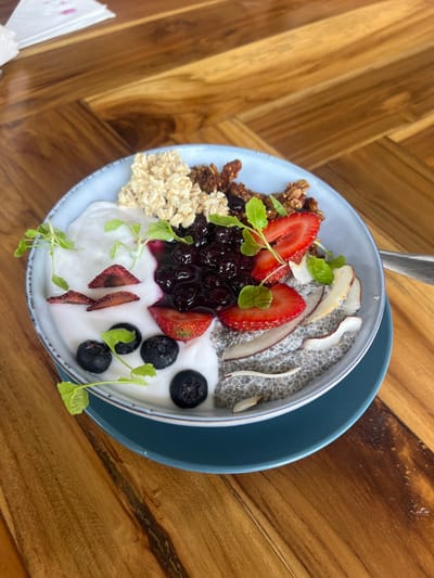A blue bowl filled with a breakfast parfait sits on a blue plate on top of a wooden table. The parfait contains yogurt, granola, blueberries, chia seeds, coconut flakes, and strawberries. The bowl is positioned in the center of the frame, with the table taking up the majority of the background. The table has a light brown wooden grain and is likely situated outdoors, as evidenced by the natural light casting shadows on the table. The setting appears to be a café or restaurant. There are no visible people in the image. The location is near a bakery in Santa Fe, Costa Rica, located at 9.642 latitude and -85.165 longitude.