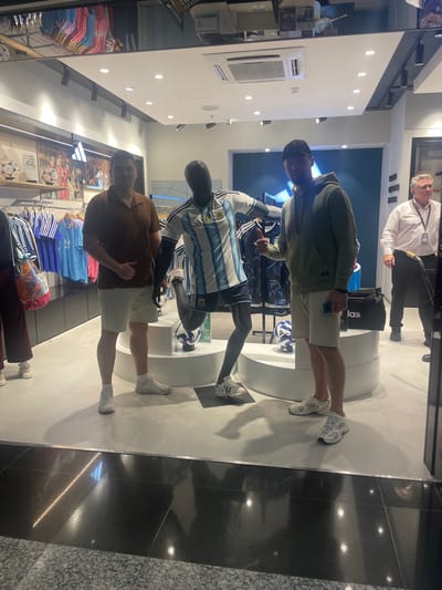 In a brightly lit Adidas store at Aeropuerto Internacional Ezeiza, Argentina, two individuals stand among displays of athletic apparel. The first individual, wearing a dark hoodie and a black baseball cap, is captured from the chest down, holding a light blue smartphone. They are standing on a white floor. Next to them, a mannequin is dressed in a blue and white striped football jersey, possibly an Argentina national team kit, with black shorts. A football rests near the mannequin.

Further down, a second individual, wearing a brown polo shirt and light-colored shorts, stands with their right hand resting on their hip. They are looking towards the camera with a relaxed expression. They are also standing on a white floor. Behind them, racks of colorful t-shirts are visible, along with framed images of athletes and footballs displayed on the wall. The store features a modern design with white walls and subtle lighting, characteristic of a retail environment in an airport. The presence of Adidas branding on a black bag and various apparel suggests a sports-focused retail space. The time of day is not explicitly indicated, but the interior lighting suggests it is daytime or the store is well-illuminated.