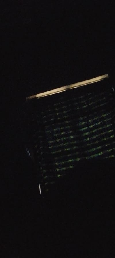 In a dimly lit or nighttime scene, a rectangular object with horizontal lines is visible. The object is dark, with subtle green highlights across the horizontal lines. A bright, linear light source illuminates the top edge of the object, creating a strong contrast with the surrounding darkness. An additional, faint light streak is visible on the left side, adding to the low-light ambience.