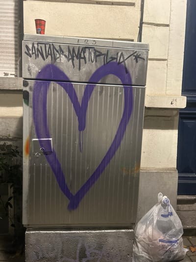 An external view in Saint-Gilles - Sint-Gillis, Belgium, depicts a tall, silver-colored, corrugated utility cabinet positioned against a light-colored building facade. The top section of the cabinet is marked with black spray-painted graffiti reading "SANTADRAMATOPILIA" and "123". Below this, a large, purple spray-painted heart shape with visible drips covers the majority of the cabinet's front. A red and black plaid disposable cup rests on the top surface of the cabinet. To the immediate right of the cabinet, a clear plastic trash bag, knotted at the top and containing refuse, sits on the ground. The building wall behind the cabinet is off-white with visible stone molding, and a dark blue door frame is partially visible to the far right. Dark green foliage is present at the base of the cabinet on its left side. The ground surface appears to be paved, with a few fallen leaves visible. No individuals are present.