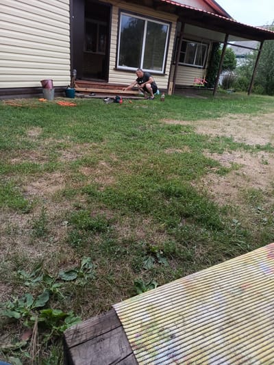A man is working on the porch of a house in the city of Vylkovo, Russia. He is wearing a black t-shirt and shorts and is crouching down to work on some wood. There are tools and buckets around him, as well as some orange extension cords. The house has light-colored siding and a dark-colored door. There is a porch with a red roof extending from the front of the house. The ground is covered in grass and dirt, and there are some trees in the background. A child's swing set is visible on the porch.