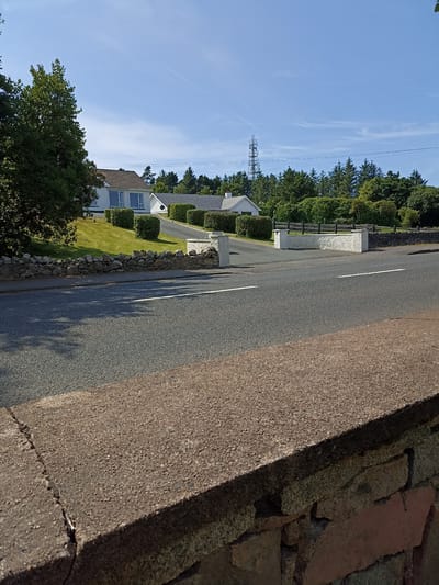 The image shows a roadside scene in Dungloe, Ireland, under a clear blue sky with wispy clouds. In the foreground, a rough-textured stone wall runs along the edge of a paved road. The road has white lines marking the lanes. Behind the wall and road, a well-maintained lawn slopes up towards two white houses with tiled roofs. The houses are bordered by neatly trimmed hedges. A driveway leads up to one of the houses. In the background, a dense line of trees forms a natural boundary. A tall communication tower is visible in the distance, silhouetted against the sky. The overall impression is of a rural or semi-rural residential area.