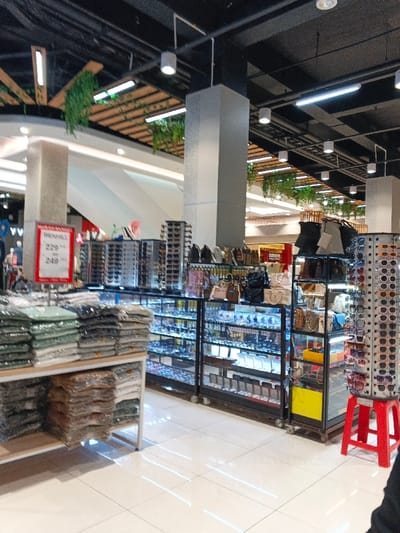 An indoor retail space features clothing on a low shelf, with several cases of sunglasses, jewelry, watches, and purses behind it. The store is brightly lit with overhead lights, and the ceiling structure is visible. A red stool stands near one of the displays. A large white support column and indoor plants are in the background.